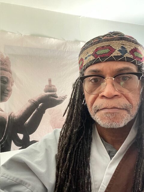 Eric Wilkins dressed in robes in front of a statue of Buddha.  A white cloth hangs in the background.