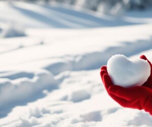 A snow-covered landscape. A person stands out of frame, red knit gloved hands visible holding a snowball shaped like a heart.